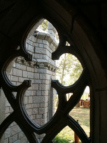 Image shows a portion of a gray masonry castle like structure as seen through open black ironwork in the shape of a cross on a sunny day